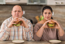 Displeased fat couple in the kitchen holding hamburgers and looking at the camera.